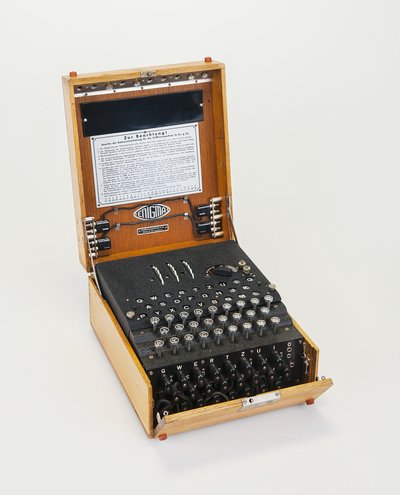 Die schwarze Maschine sitzt in einem hölzernen Kasten, der aufgeklappt ist. Oben hat sie eine Buchstabentastatur eine Buchstabenanzeige sowie drei Drehrädchen. An der Vorderseite der Maschine befinden sich schwarze doppelpolige Buchsen.