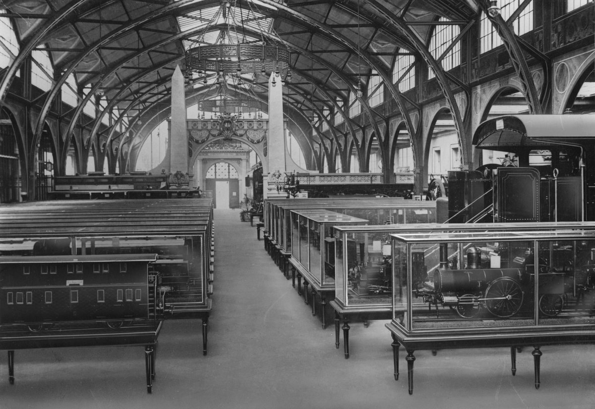 Schwarzweißfoto: Die Ausstellung in der Industriehalle des ehemaligen Hamburger Bahnhofs. Unter der aufwendigen Dachkonstruktion aus Stahl und Glas ist der Raum mit Vitrinen gefüllt, in denen Eisenbahnmodelle zu erkennen sind.