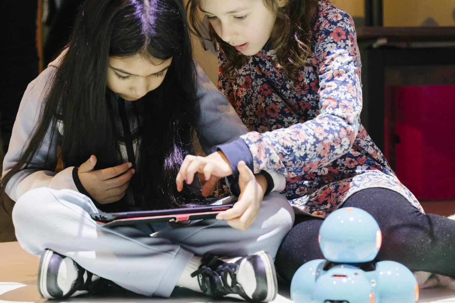 Zwei Mädche knien neben einem kleinen blauen Roboter. Ein Mädchen hält ein tablet in den Händen, das zweite Mädchen zeigt ihr etwas darauf.