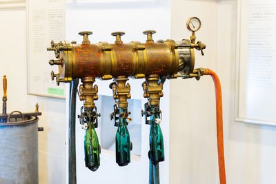 Drei Flaschen sind in die zylinderförmige Maschine zur Befüllung eingehängt. Ein Schlauch führt zur Befüllungsmaschine, über dem Schlauch ist ein Druckmesser mit Anzeige angebracht.