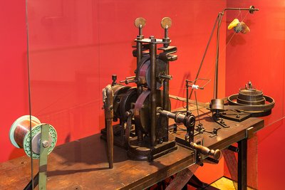 Die Strickmaschine aus Metall steht in der Mitte einer hölzernen Werkbank. An der Vorderseite sitzt eine Handkurbel. Links der Maschine steht eine weiße Spindel. Ein Metall-Faden wird durch die beiden übereinanderliegenden Räder der Maschine geführt.