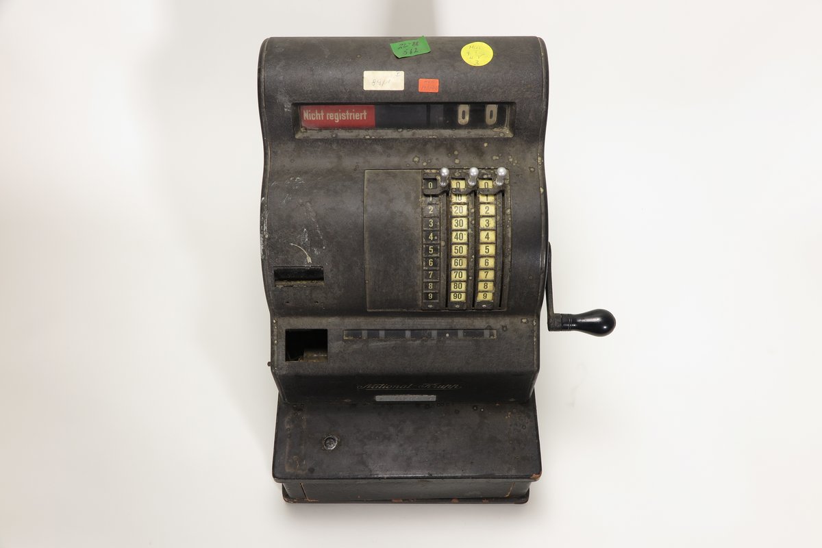 Registrierkasse aus den 1930er Jahren mit vier farbigen Aufklebern der KuA. Die Aufkleber tragen Zahlen und Buchstaben.