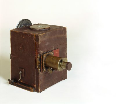 Die Kamera ist ein einfacher brauner Holzkasten. An der Vorderseite sitzt das Objektiv, an der rechten Seite befindet sich eine kleine Handkurbel.