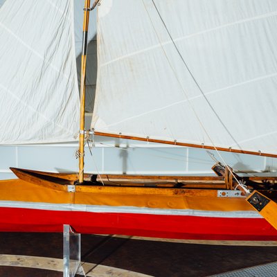 Ein langes Sportsegelboot hängt über dem Holzboden. Es hat drei aufgespannte weiße Segel und einen rotbraunen Rumpf. Im Hintergrund ragt ein weiteres Segel aus weißen, horizontal gespannten Wellenbändern hervor.