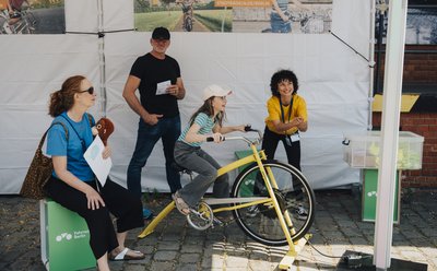 Mehrer Menschen stehen um ein fest montiertes Fahrrad herum, ein Mädchen fährt darauf.