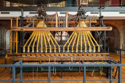 Der Webstuhl ist etwa fünf Meter breit, 2,4 Meter tief und 3,5 Meter hoch. Die beiden Webstuhlaufsätze sind oben an einem Metallrahmen aufgehängt. Sie sehen aus wie zwei riesige Rechen mit je acht Zähnen.