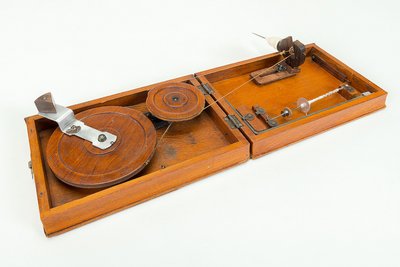 In einem aufgeklappten, flachen Holzkasten sieht man rechts die Spindel mit einem Baumwollfaden und links das kleine Spinnrad, das über einem Riemen mit dem größeren Antriebsrad verbunden ist. Auf dem Antriebsrad sitzt ein Metallgriff mit einem Holzknauf.