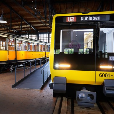 Blick auf zwei nebeneinander stehende U-Bahnwagen. Beide sind gelb. Der Wagen rechts sieht sehr modern aus, der Wagen links ist ein historisches Fahrzeug.