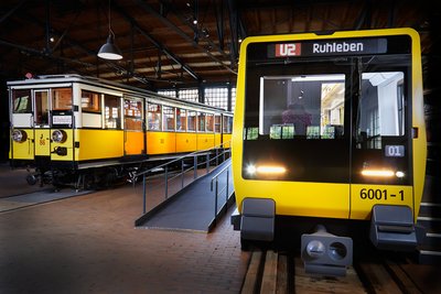 Blick auf zwei nebeneinander stehende U-Bahnwagen. Beide sind gelb. Der Wagen rechts sieht sehr modern aus, der Wagen links ist ein historisches Fahrzeug.