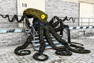 Der Datenkrake Otto liegt auf einer breiten Metalltreppe vor einem Holztor. Seine acht schwarzen Arme sind über die Stufen drapiert. Sein Auge leuchtet gelb. Auf Kopf und Armen sind viele gelbe Nullen und Einsen gemalt.