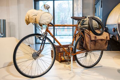 Das Sportrennrad Dinos hat einen orangenen Rahmen. Am Lenker ist über dem Vorderrad eine große weiße Packtasche aus Stoff angebracht. Zu beiden Seiten des Hinterrads sitzen zwei braune Packtaschen. Auf dem Gepäckträger sind ein Wäschesack und zwei Ersatzschläuche befestigt.