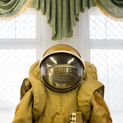 Das Bild zeigt zentral einen Kosmonautenanzug von der Taille aufwärts. Darauf sitzt ein kugelförmiger Helm mit großem Glasvisier. Im Hintergrund hängt über einem Fenster eine Zierschabracke mit Goldbordüre.