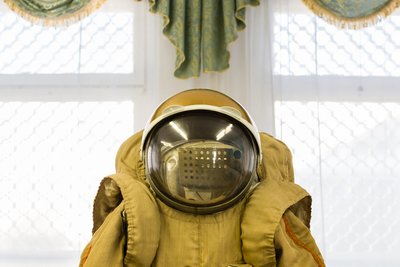 Das Bild zeigt zentral einen Kosmonautenanzug von der Taille aufwärts. Darauf sitzt ein kugelförmiger Helm mit großem Glasvisier. Im Hintergrund hängt über einem Fenster eine Zierschabracke mit Goldbordüre.