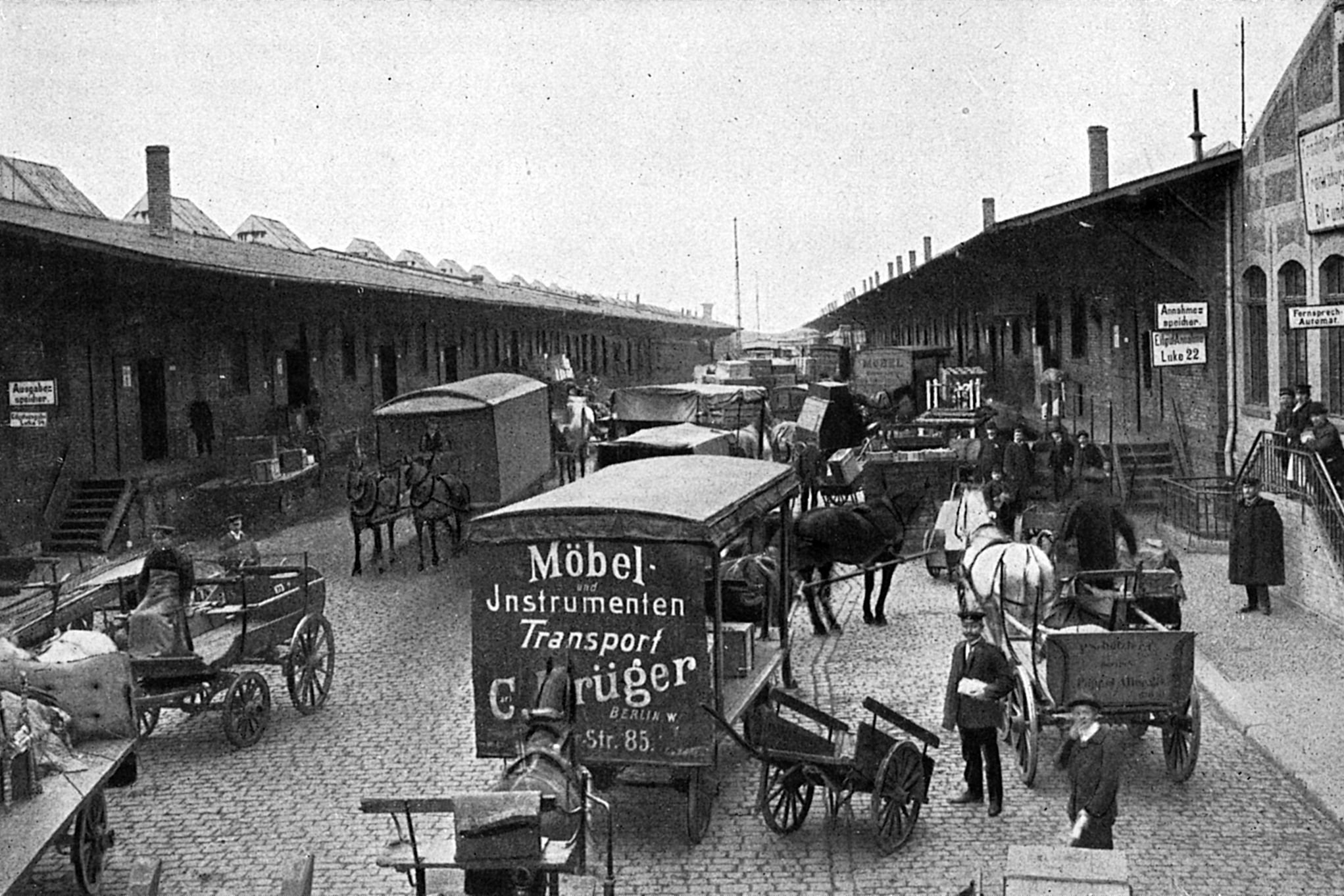Schwarzweiß-Foto einer kopfsteingepflasterten Straße mit Lagerschuppen auf beiden Seiten. Auf der Straße sind Pferdefuhrwerke, Karren und Menschen unterwegs.