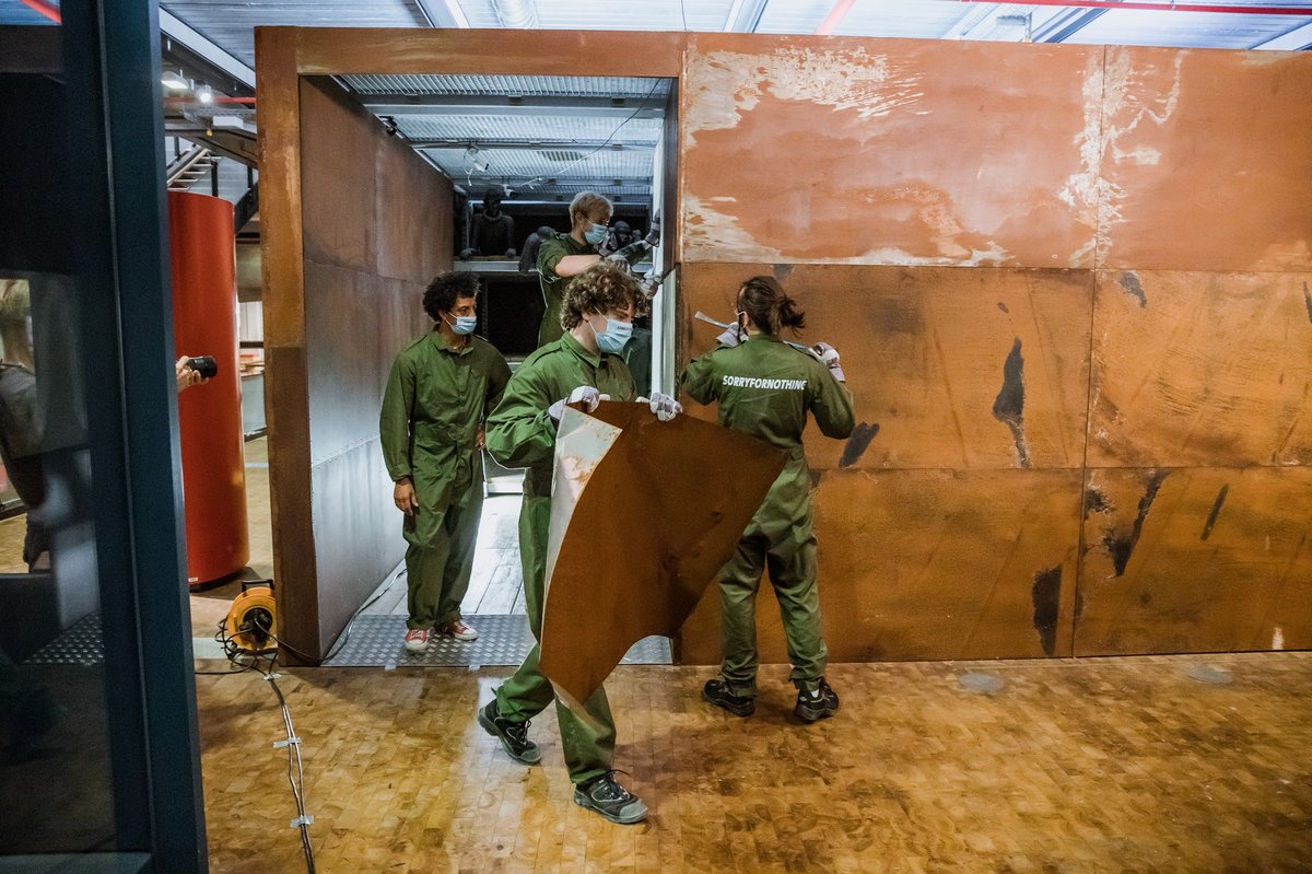 Mehrere Menschen in grünen Arbeitsanzügen transportieren Materialien aus einem braunen Metallkubus.