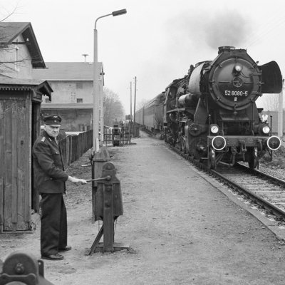 Eine Schwarzweiß-Fotografie: Auf einem Bahnsteig in ländlicher Umgebung steht ein älterer Schrankenkwärter in Uniform. Von hinten nähert sich eine Dampflokomotive.