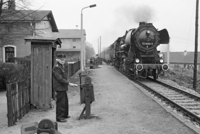 Eine Schwarzweiß-Fotografie: Auf einem Bahnsteig in ländlicher Umgebung steht ein älterer Schrankenkwärter in Uniform. Von hinten nähert sich eine Dampflokomotive.
