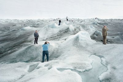 Eine Gruppe von Touristinnen und Touristen steht auf einer schmelzenden Eislandschaft und posiert an verschiedenen Punkten des Bildes für Fotos. An vielen Stellen der Eislandschaft hat sich schwarzer Ruß abgesetzt, das Eis ist dort sehr dunkel. Im Vordergrund ist eine große Pfütze aus Schmelzwasser.