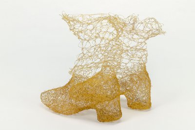 Zwei Stiefel stehen nebeneinander. Sie sind aus feinem Kupferdraht gewebt, der wie ein feiner Goldfaden aussieht. An den Füßen und den Hacken ist das Material dichter gewoben. Oben wird das Material luftiger, als würden die Stiefel sich auflösen.
