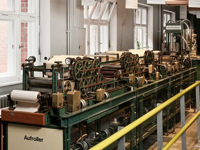 Die Maschine ähnelt einer mehrere Meter langen Werkbank. Auf ihr sitzt ein System von mehr als 15 Walzen verschiedener Größen, durch die die Papierbahn läuft.