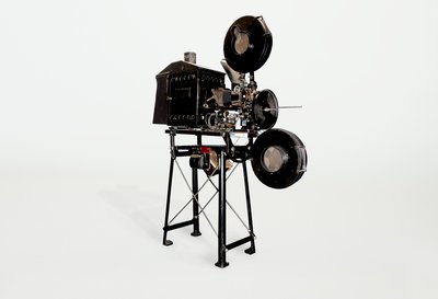 Der Filmprojektor Ernemann I ist auf einem Vorführtisch montiert. Oben über dem Objektiv und schräg nach vorne versetzt, unter dem Objektiv, sind zwei Filmrollen angebracht.