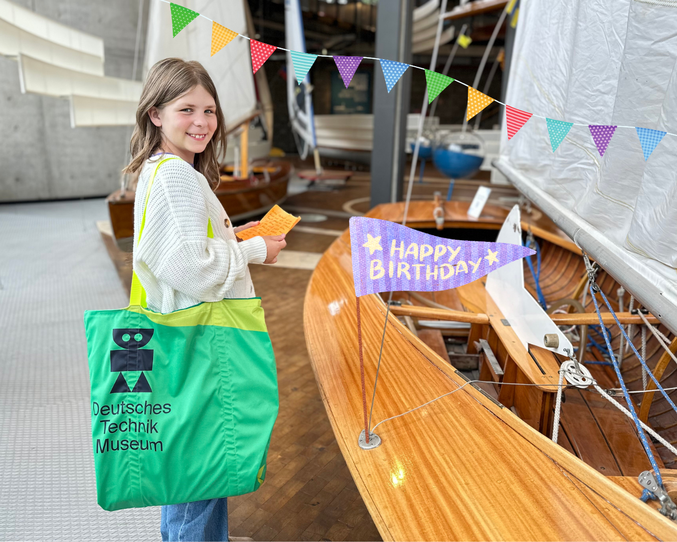 Eine junge Person seht neben einem historischen Segelboot aus Holz mit einem weißen Segel in einem Museum. Die person hat eine grüne Tasche mit dem Logo des Museums und dem Schriftzug "Deutsches Technikmuseum" über dei rechte Schulter gehängt und lächelt.