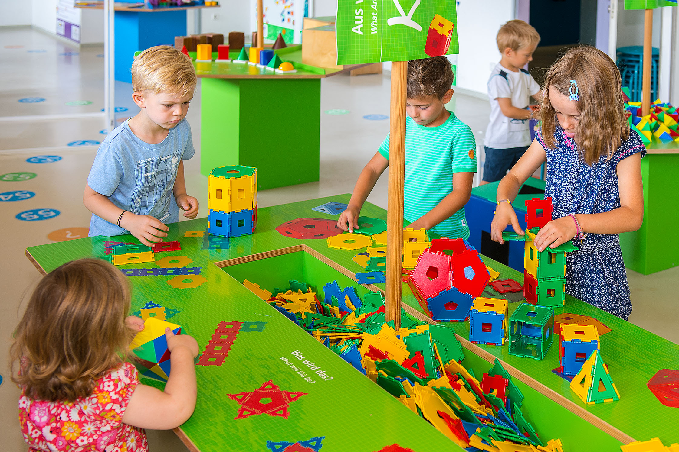 Eine Gruppe von Kindern steht um einen grünen Tisch herum, auf dem verschiedene geometrische Formen zum Zusammenbasteln liegen.