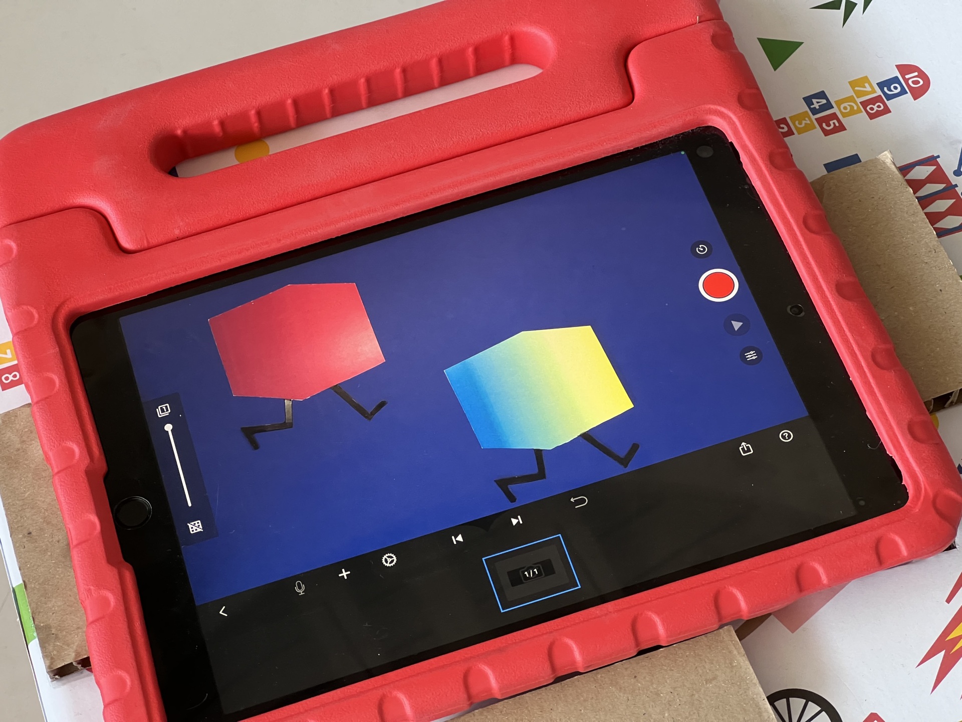 Tablet in roter Schutzhülle mit geöffneter App „Stop Motion Studio"; auf dem Bildschirm sind zwei farbige Figuren mit beweglichen Beinen zu sehen.