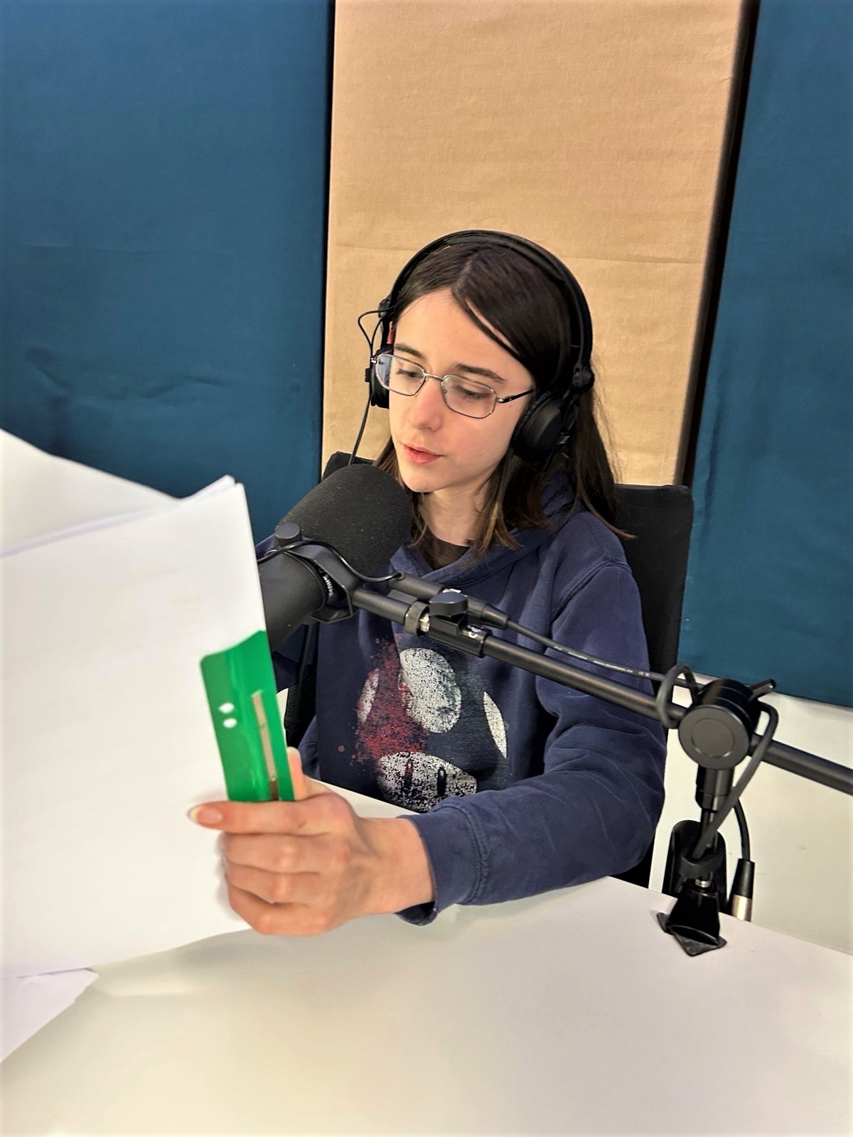 Eine junge Person mit braunen schulterlangen Haaren und einer Brille sitzt in einem Tonstudio vor einem Mikrofon und hält ein Skript in der Hand.