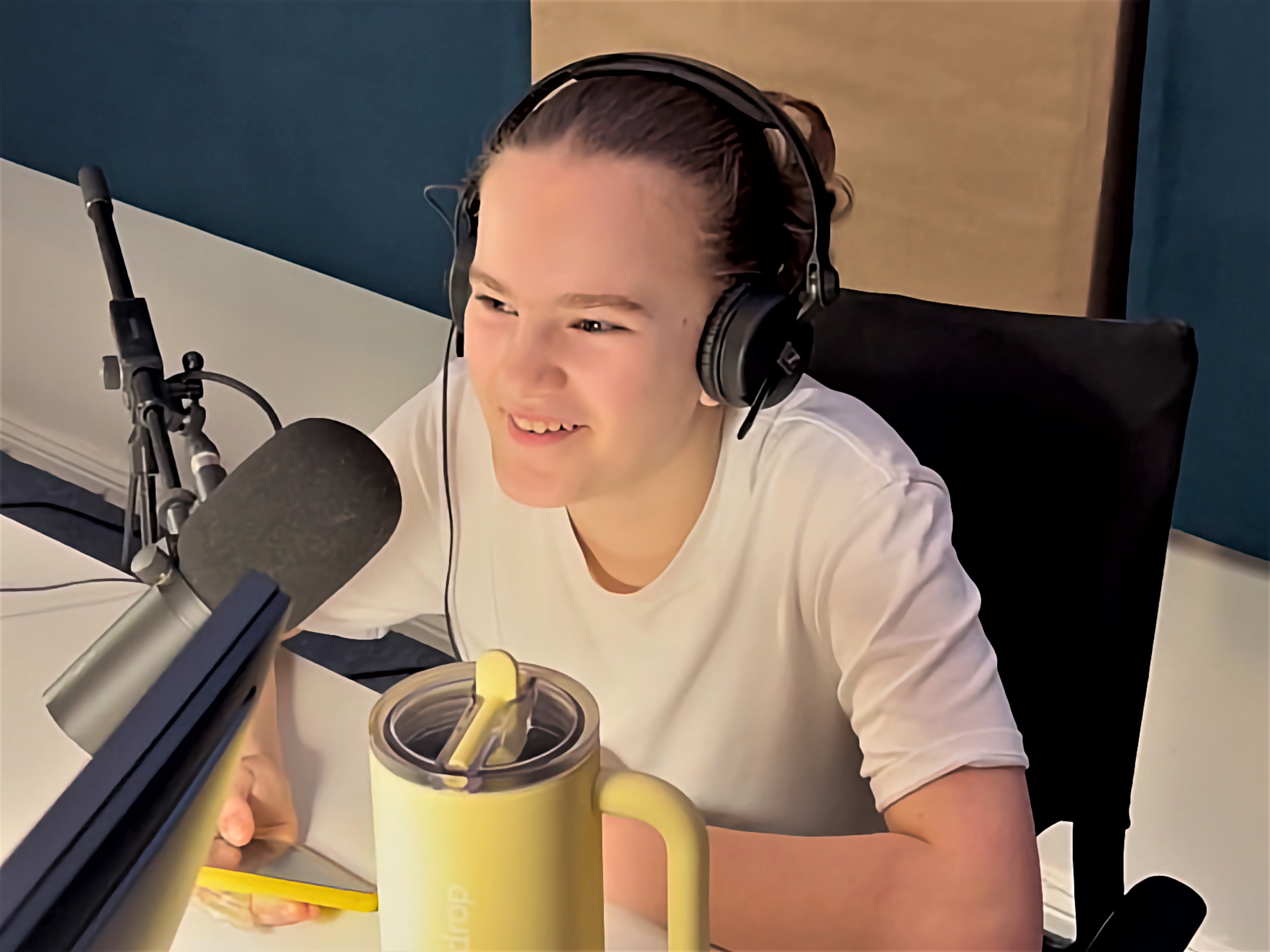 Eine junge Person mit braunen Haaren sitzt in einem Tonstudio vor einem Mikrofon und lächelt. Die person hält ein Smartphone mit einer gelben Schutzhülle in der rechten Hand.