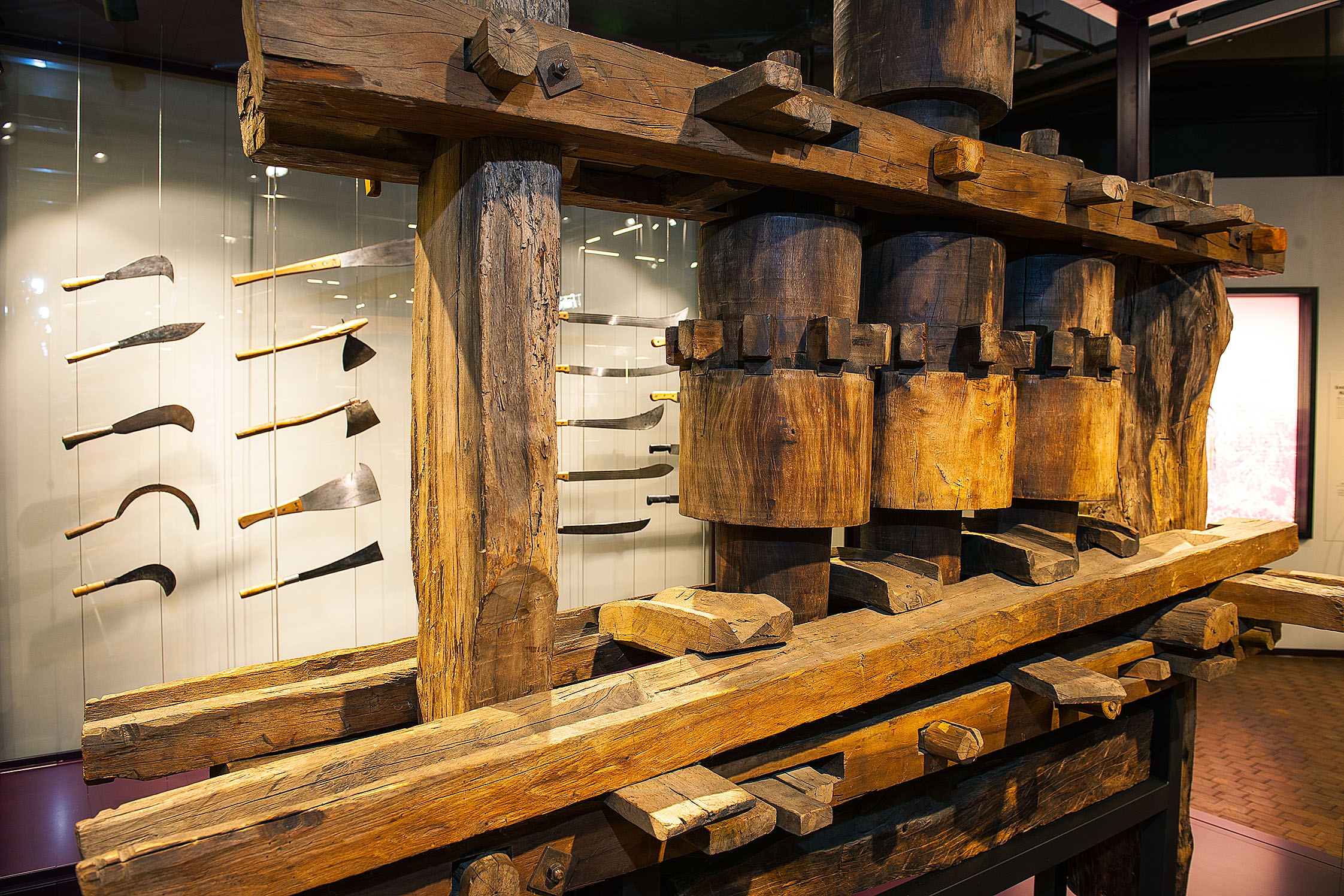 Eine große, hölzerne Zuckerrohrmühle. Sie besteht aus einer langen Antriebsstange oben, die eine Antriebswalze zum Rotieren bringt. Diese setzt zwei weitere Walzen in Gang, die das Zuckerrohr auspressen.