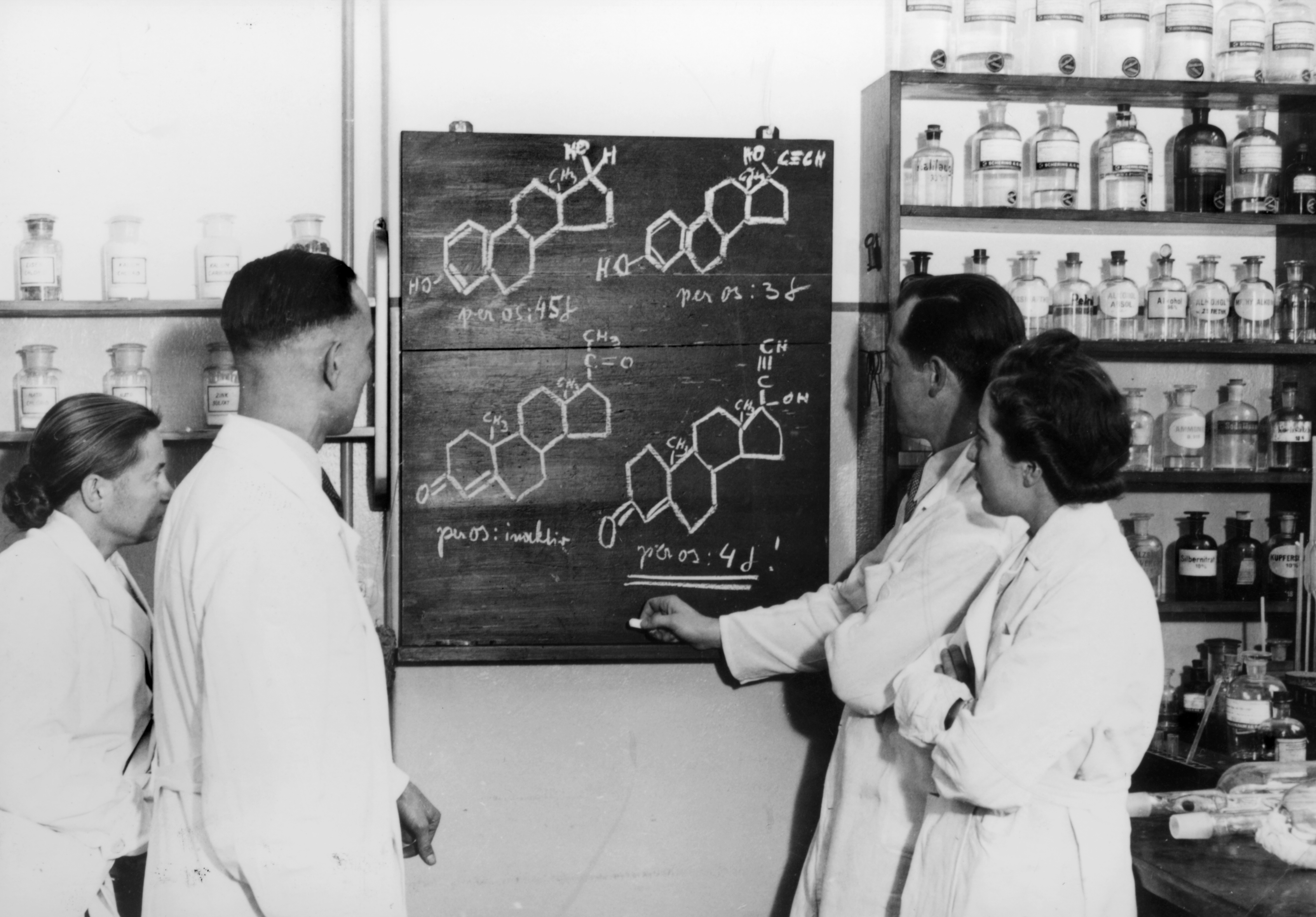 Schwarzweißfoto, 1938: Im Labor der Firma Schering stehen zwei Mitarbeiter und eine Mitarbeiterin vor einer Tafel. Auf dieser sind mit Kreide Molekularstrukturformeln gezeichnet.