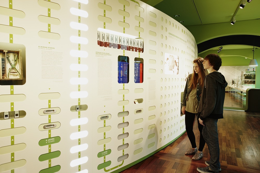 Eine Besucherin und ein Besucher betrachten eine Infografik, die mithilfe von grünen und weißen Lichtfeldern den Weg von der Wirkstoffsuche bis zum fertigen Arzneimittel verdeutlicht.