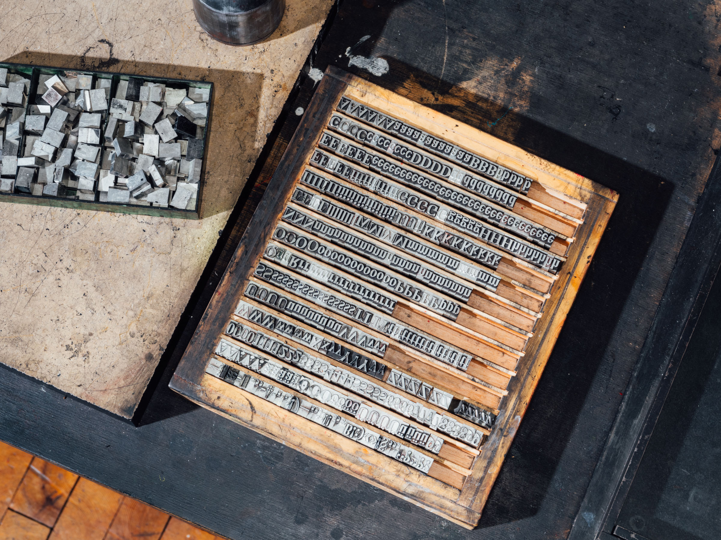 In einem Holzkasten mit einzelnen waagrechten Zeilen sind Metallettern zu Sätzen angeordnet. Daneben ein Kasten mit losen Lettern.