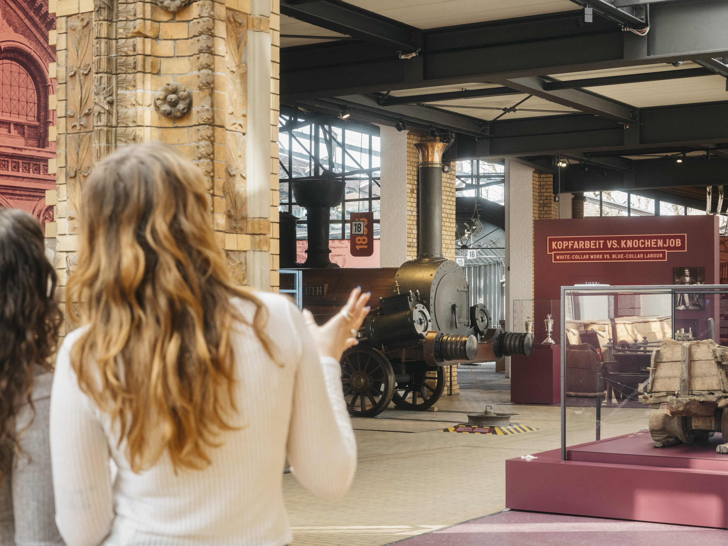 Im Vordergrund stehen zwei Frauen mit dem Rücken zur Kamera. Sie schauen auf die Ausstellung Schienenverkehr. Im Hintergrund ist ein Teil einer Lokomotive zu erkennen, dazu Vitrinen und Texttafeln.