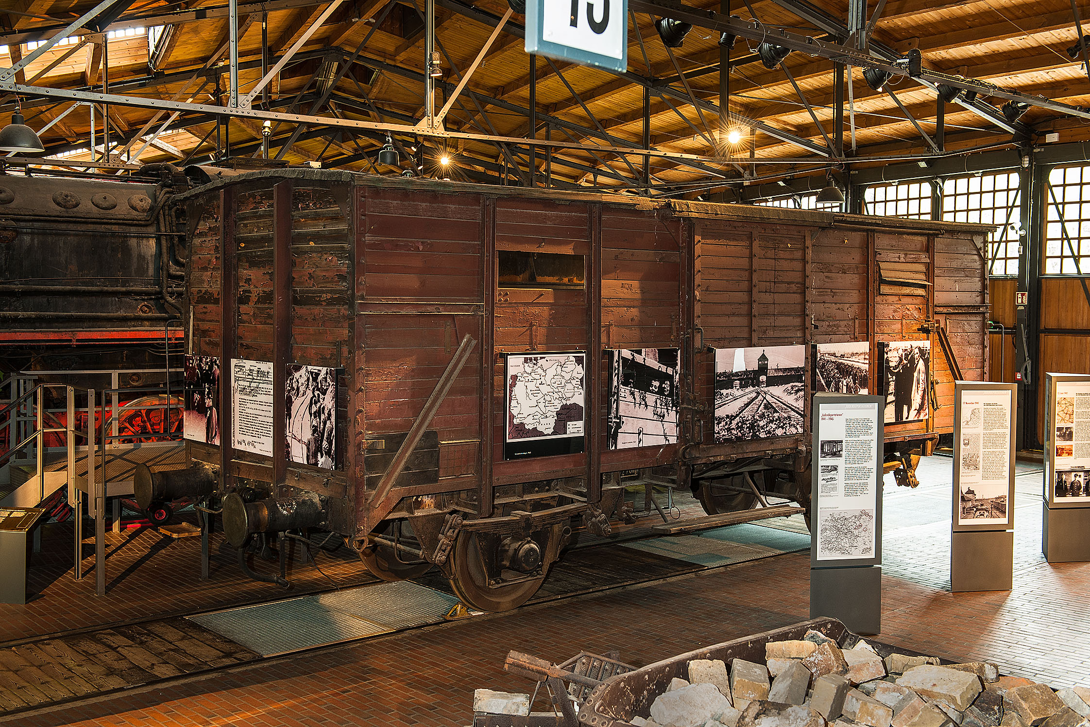 Blick auf einen alten braunen Güterwagen mit Holzaufbau. Auf den Seiten sind Schwarz-Weiß-Aufnahmen aus Konzentrationslagern zu sehen.