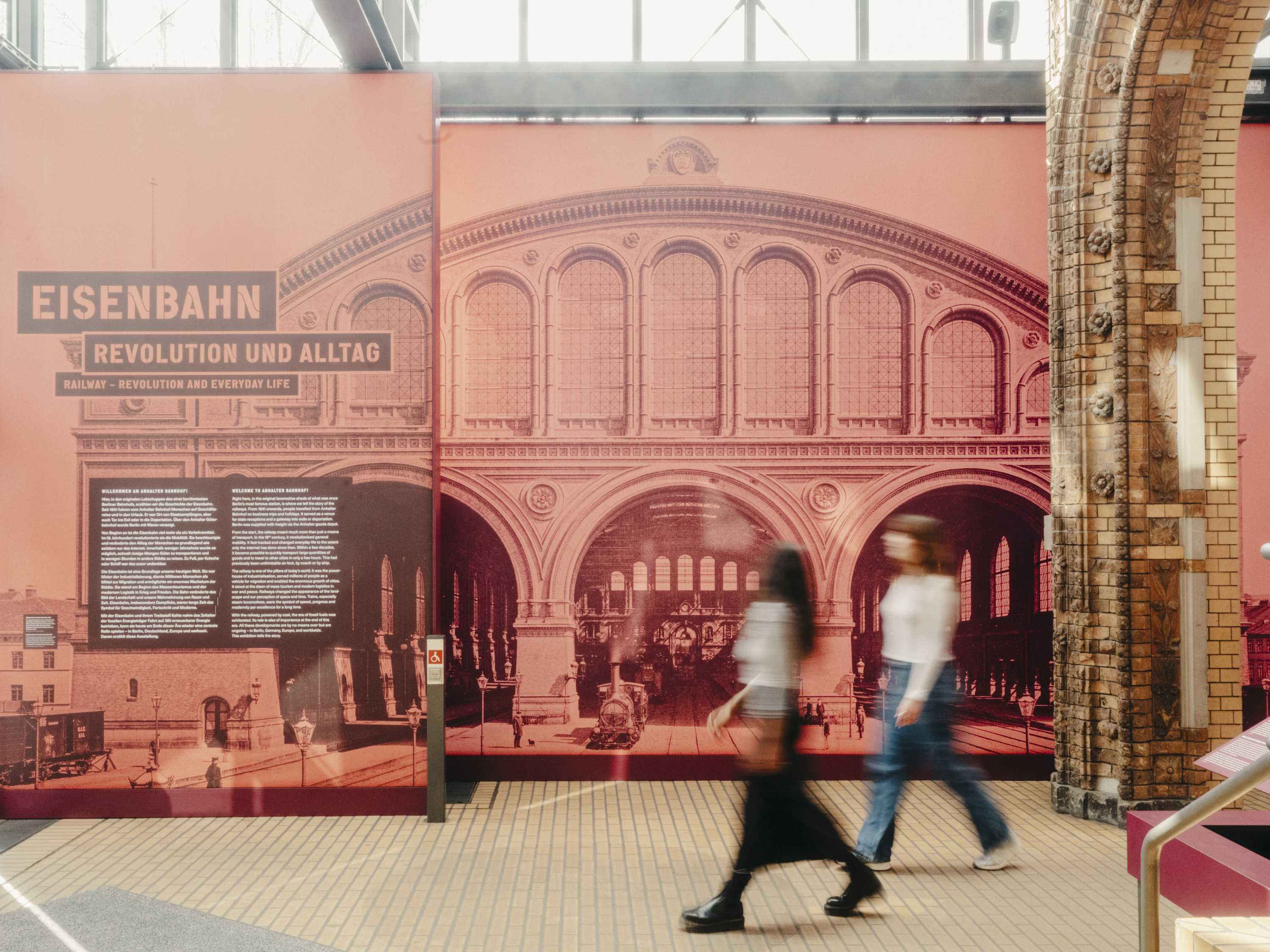 Zwei Frauen laufen von rechts nach links durch das Bild. Rechts hinter ihnen ist der Bogen eines alten Portals erkennbar. Direkt hinter ihnen befindet sich eine Wand, die mit einer rot eingefärbten Fotografie des Anhalter Bahnhofs bedruckt ist. Links in diesem Foto befindet sich ein Textfeld mit der Aufschrift: „Eisenbahn: Revolution und Alltag“.