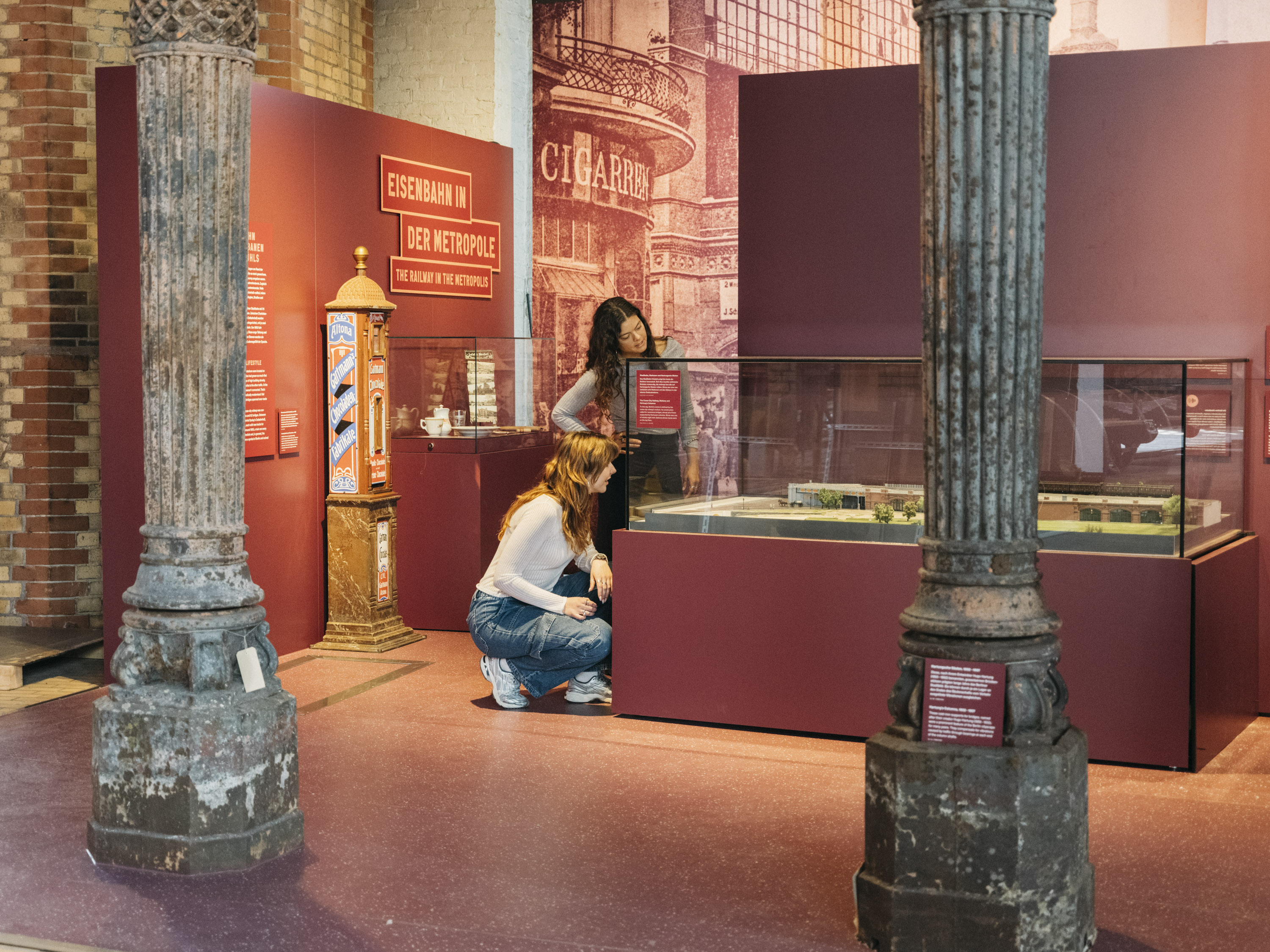 Zu sehen ist die in weinrot gehaltene Themeninsel „Eisenbahn in der Metropole“ der überarbeiteten Ausstellung Schienenverkehr im Deutschen Technikmuseum. Im Vordergrund zwei alte, verzierte Metallsäulen, die sogenannten Hartungschen Säulen. Im Hintergrund betrachten zwei Personen den Inhalt einer Vitrine.