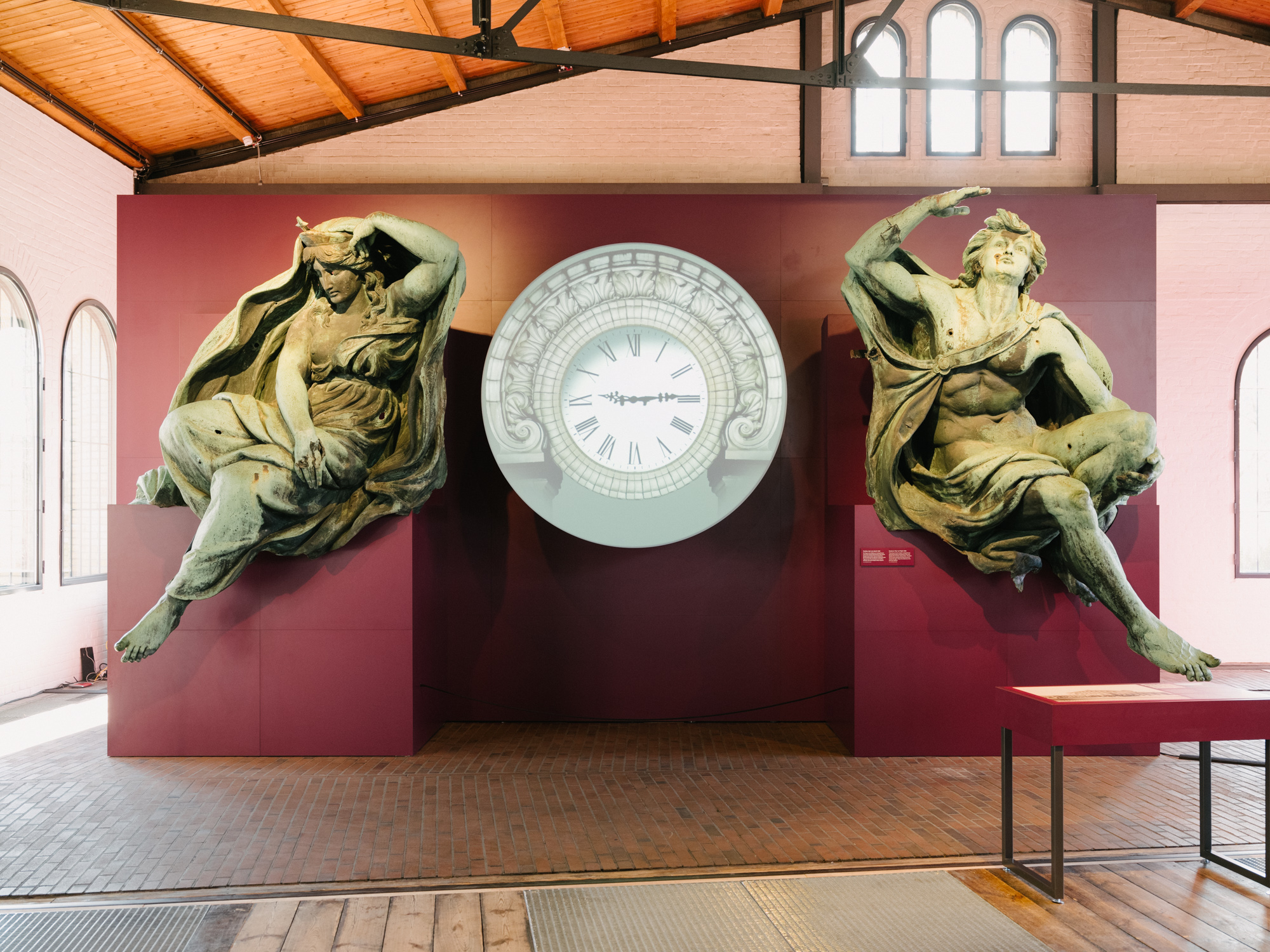 Zwei große, grün angelaufene Bronze Figuren stehen links und rechts vor einer himbeerfarbenen Wand in einem Lokschuppen. Zwischen Ihnen hängt eine Uhr.