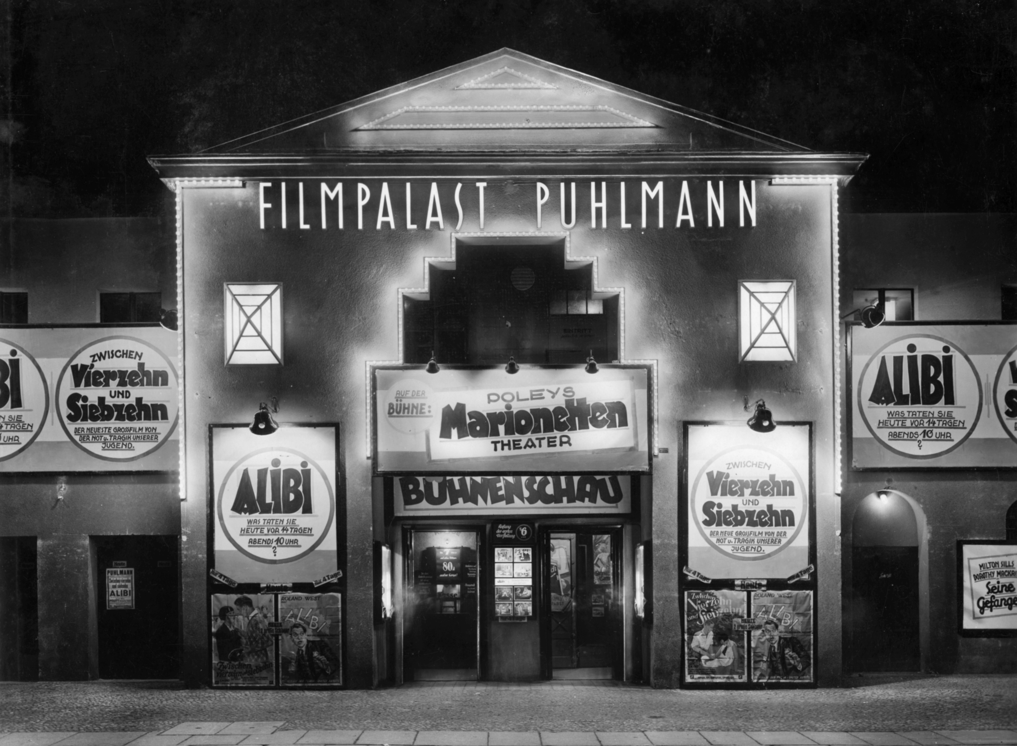 Schwarzweiß-Foto: Beleuchtetes Gebäude mit großem Schriftzug „Filmpalast Puhlmann“. An der Fassade sind viele Plakate und Banner angebracht, die für verschiedene Filme werben.