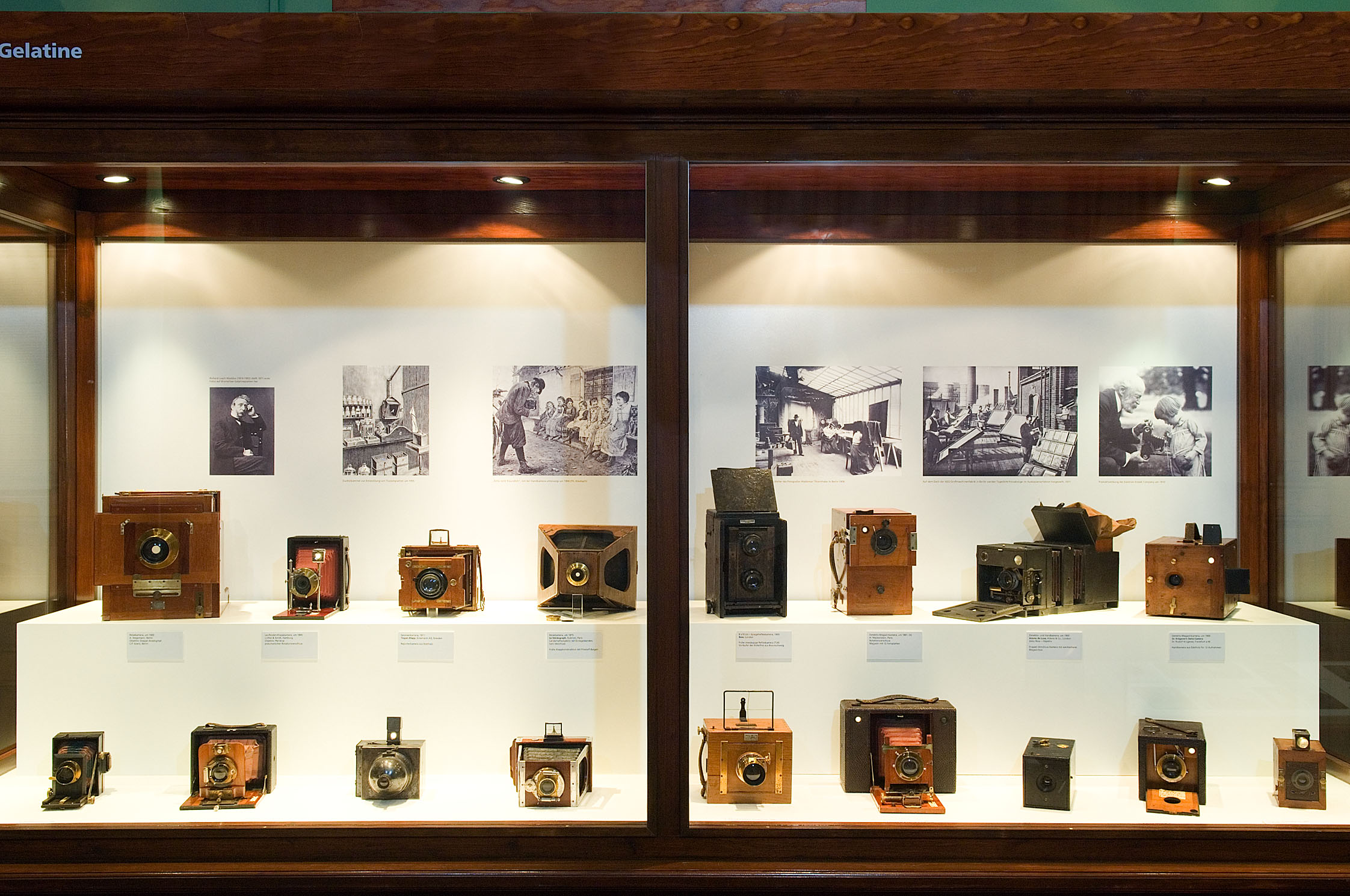 Eine große Vitrine aus dunklem Holz zeigt verschiedene historische Kameras mit Holzgehäuse. Im Hintergrund hängen einige Schwarzweißaufnahmen.