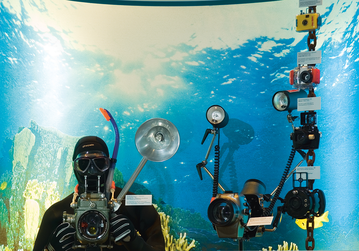 Blick in einen Schaukasten mit Unterwasseratmosphäre. Zu sehen ist ein lebensgroßer Taucher in schwarzem Taucheranzug und Taucherbrille mit Schnorchel und Unterwasserkamera in der Hand. Rechts daneben sind weitere Unterwasserkameras angebracht. 