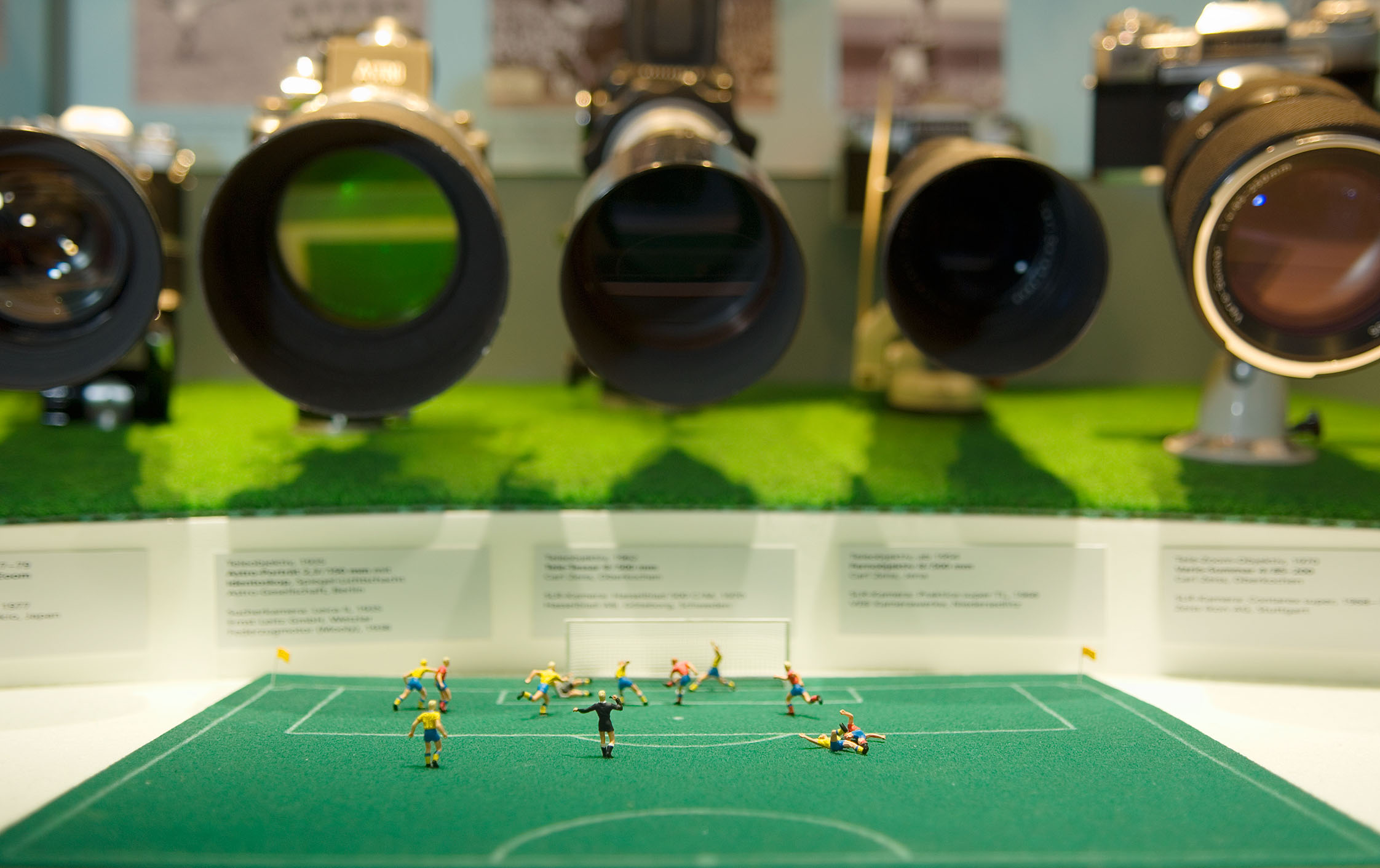Blick in eine Vitrine: Im Vordergrund liegt ein Fußballfeld aus grünem Filz, auf dem Miniaturfiguren einem Tor entgegen stürmen. Im Hintergrund sind fünf verschiedene Foto-Objektive auf die Szene gerichtet.