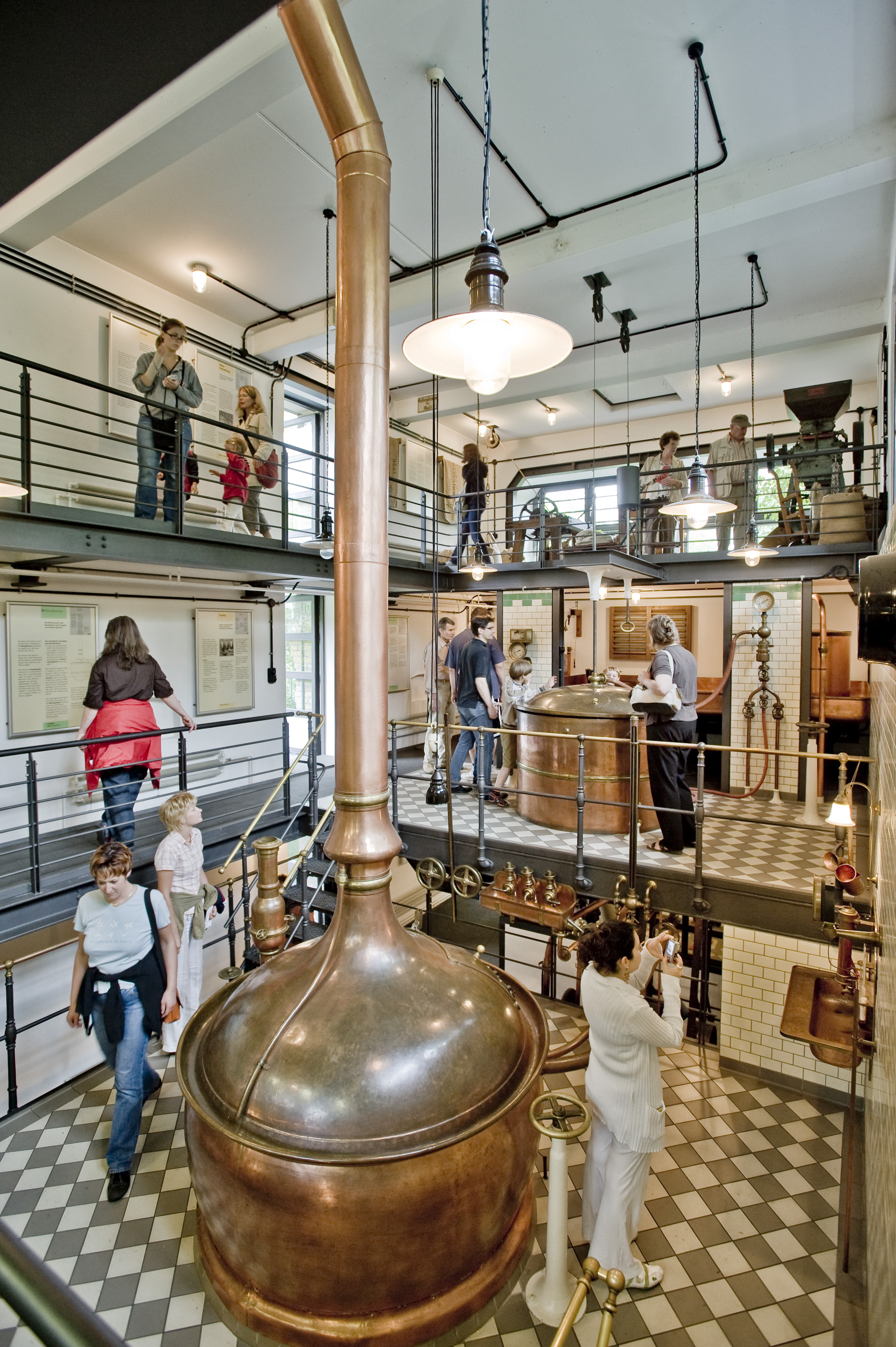 Im Zentrum der historischen Brauerei steht ein großer Bottich aus Kupfer, aus dem ein großes Rohr nach oben ragt. Besucherinnen und Besucher schauen sich auf den umliegenden zwei Etagen die Objekte in der Brauerei an.