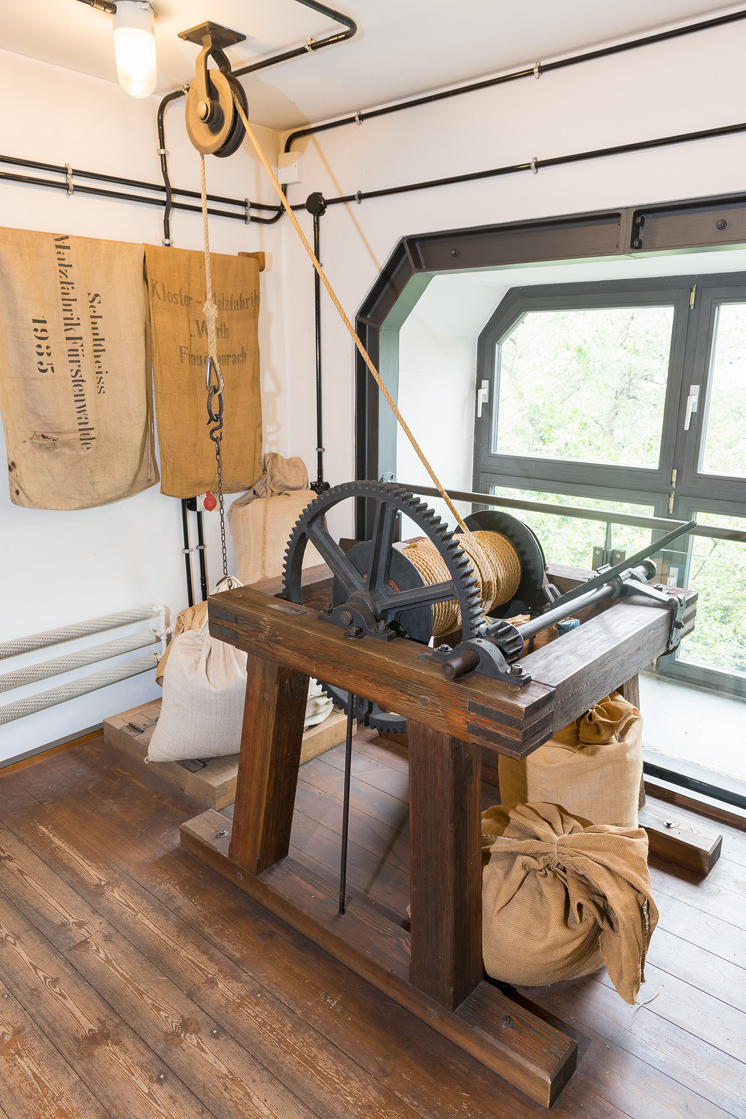 Vor einem Fenster steht ein historischer Lastenaufzug. Ein Seil läuft über einen Flaschenzug an der Decke. Um die Maschine herum und an der Wand sind historische Jutesäcke platziert.