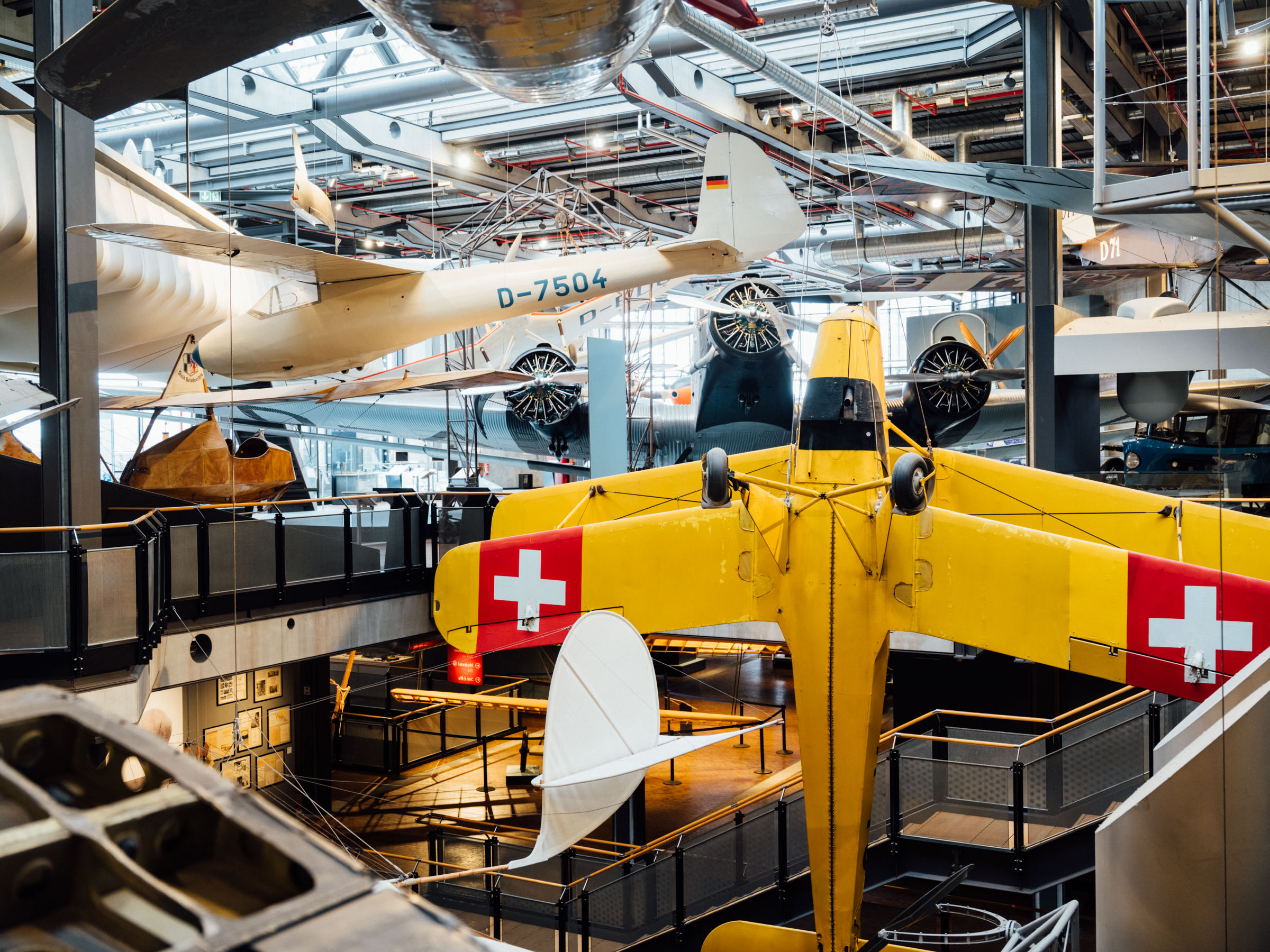 Ein großes gelbes Flugzeug hängt senkrecht im offenen Treppenhaus des Neubaus. Im Hintergrund hängen weitere Flugzeuge und Flugapparate. 