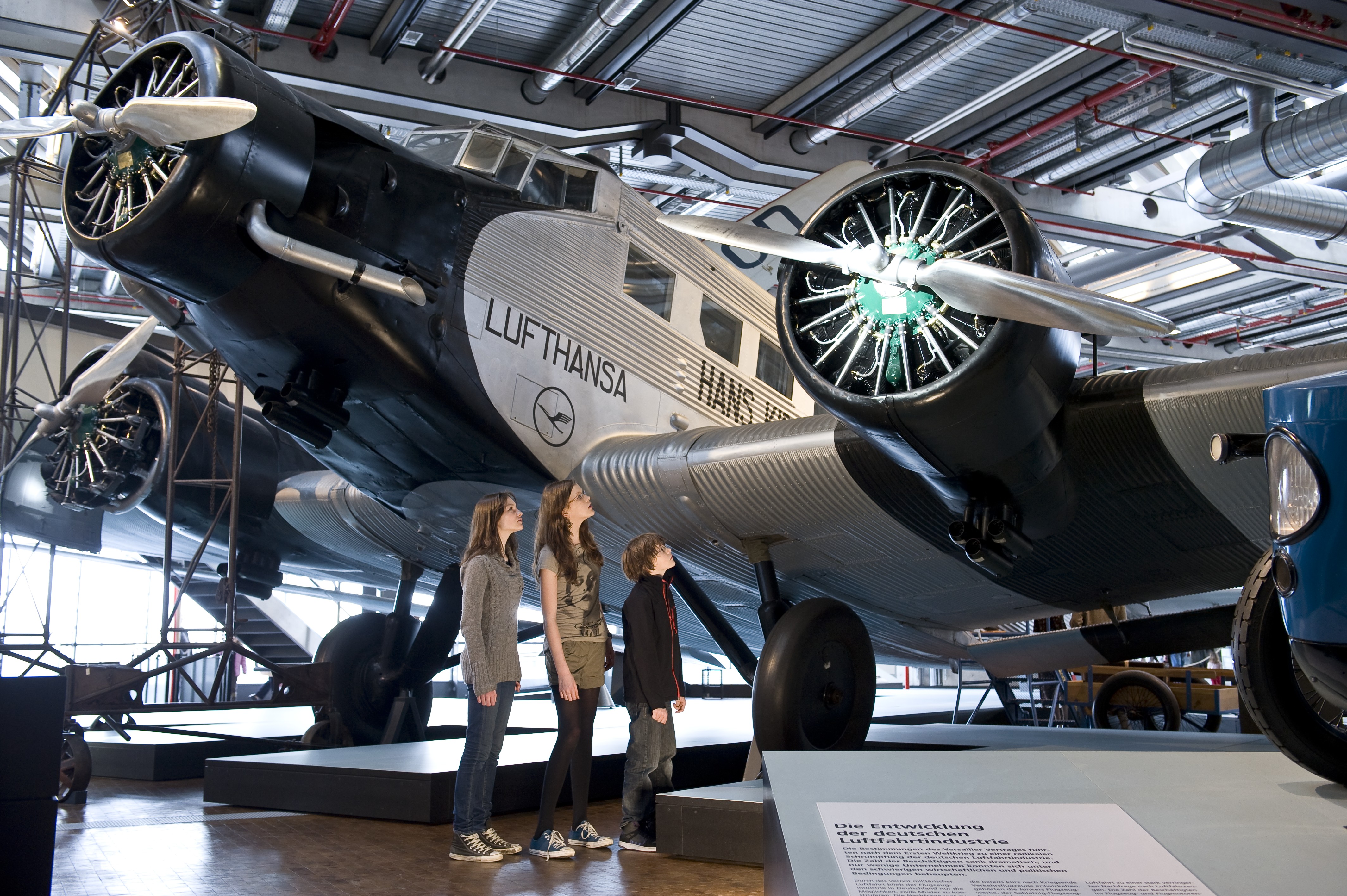 Drei Kinder stehen vor einem sehr großen Flugzeug in der Ausstellung und betrachten das Triebwerk.