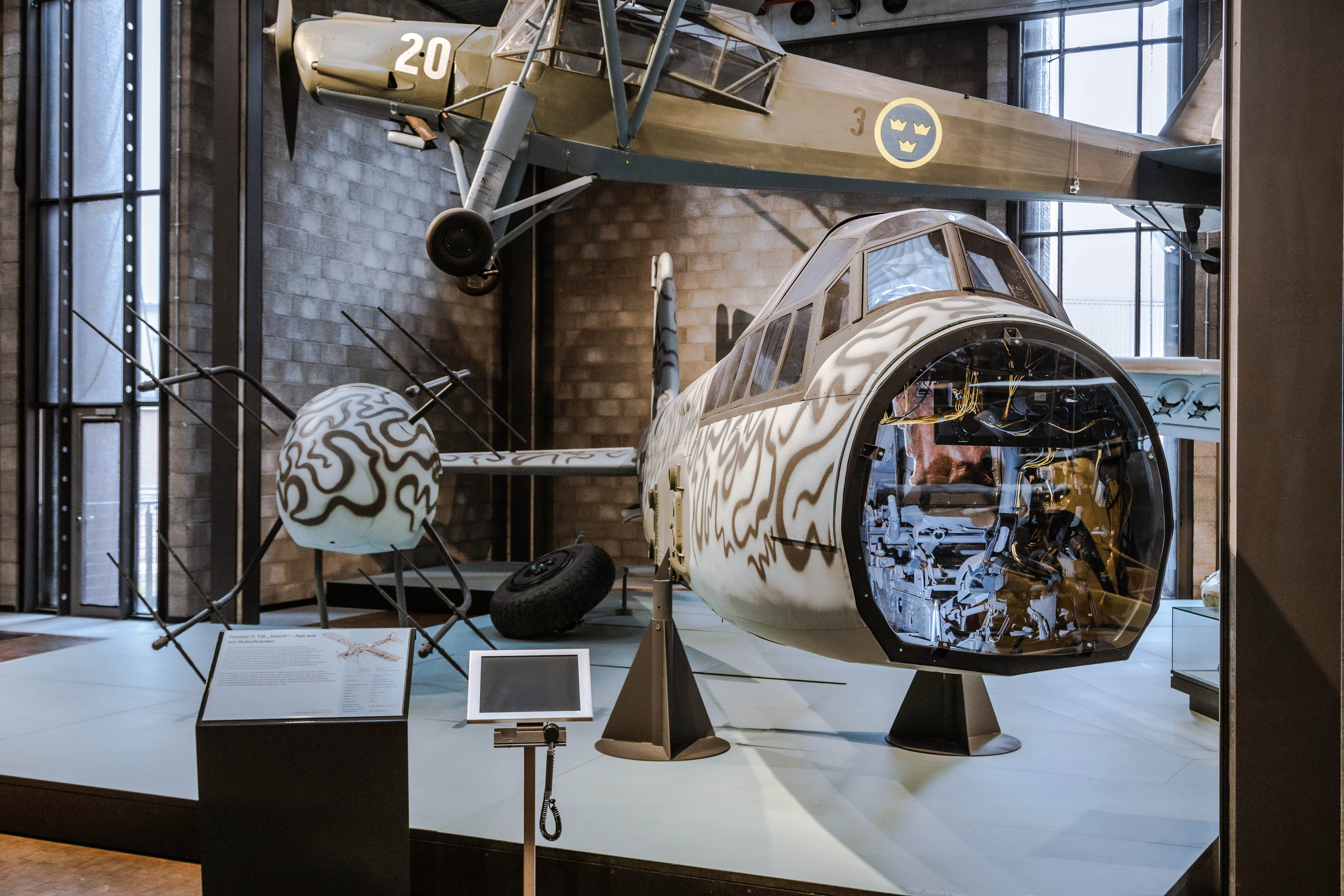Frontaler Blick auf ein Kampfflugzeug. Die Nase des Flugzeugs ist abmontiert, so dass man in das beleuchtete Innere blicken kann. Im Hintergrund hängt ein weiteres Flugzeug. 