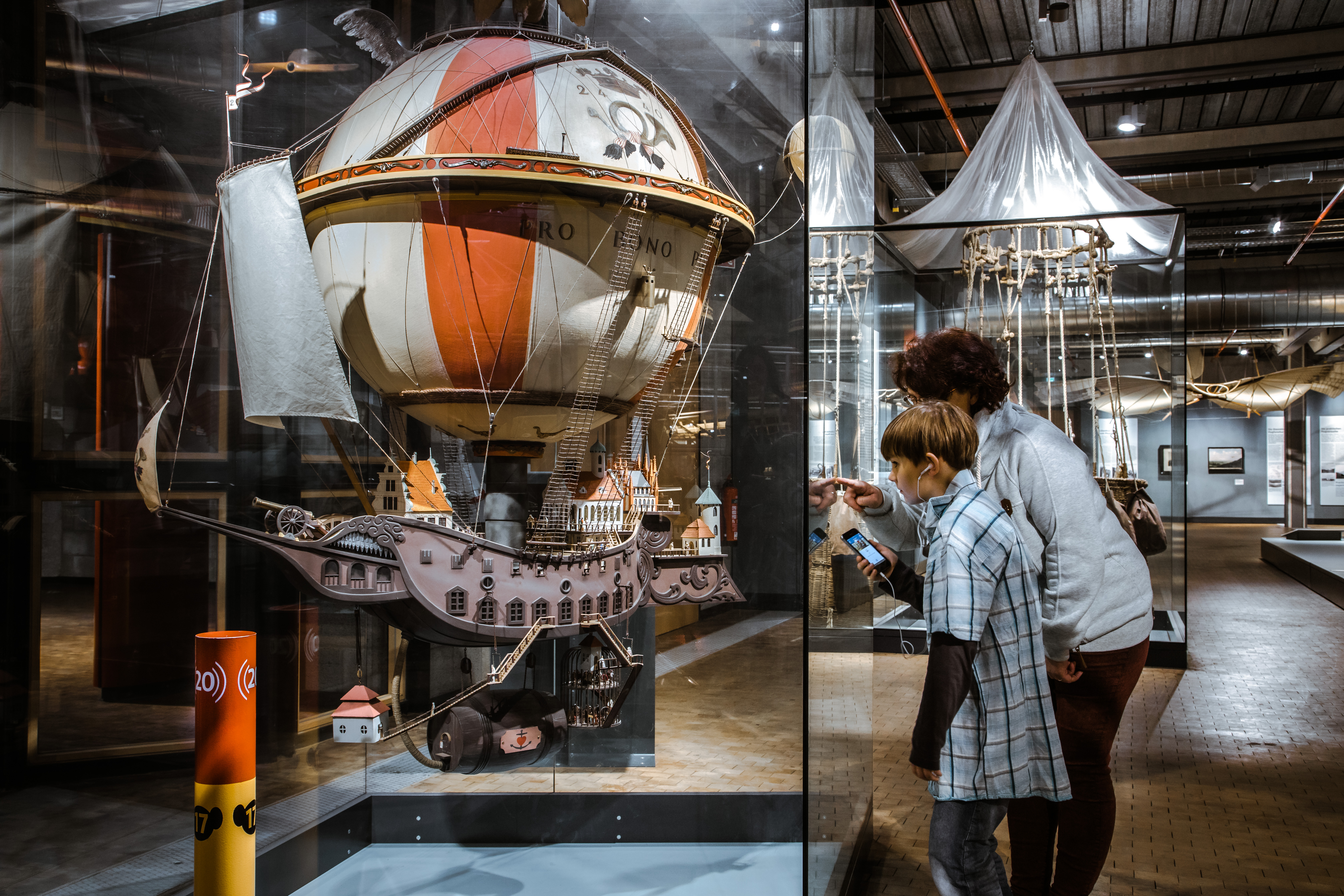 Das Modell eines großen, rotweiß gestreiften Heißluftballons an dem eine Stadt auf einem Schiffsrumpf hängt. Eine Frau und ein Kind betrachten es.