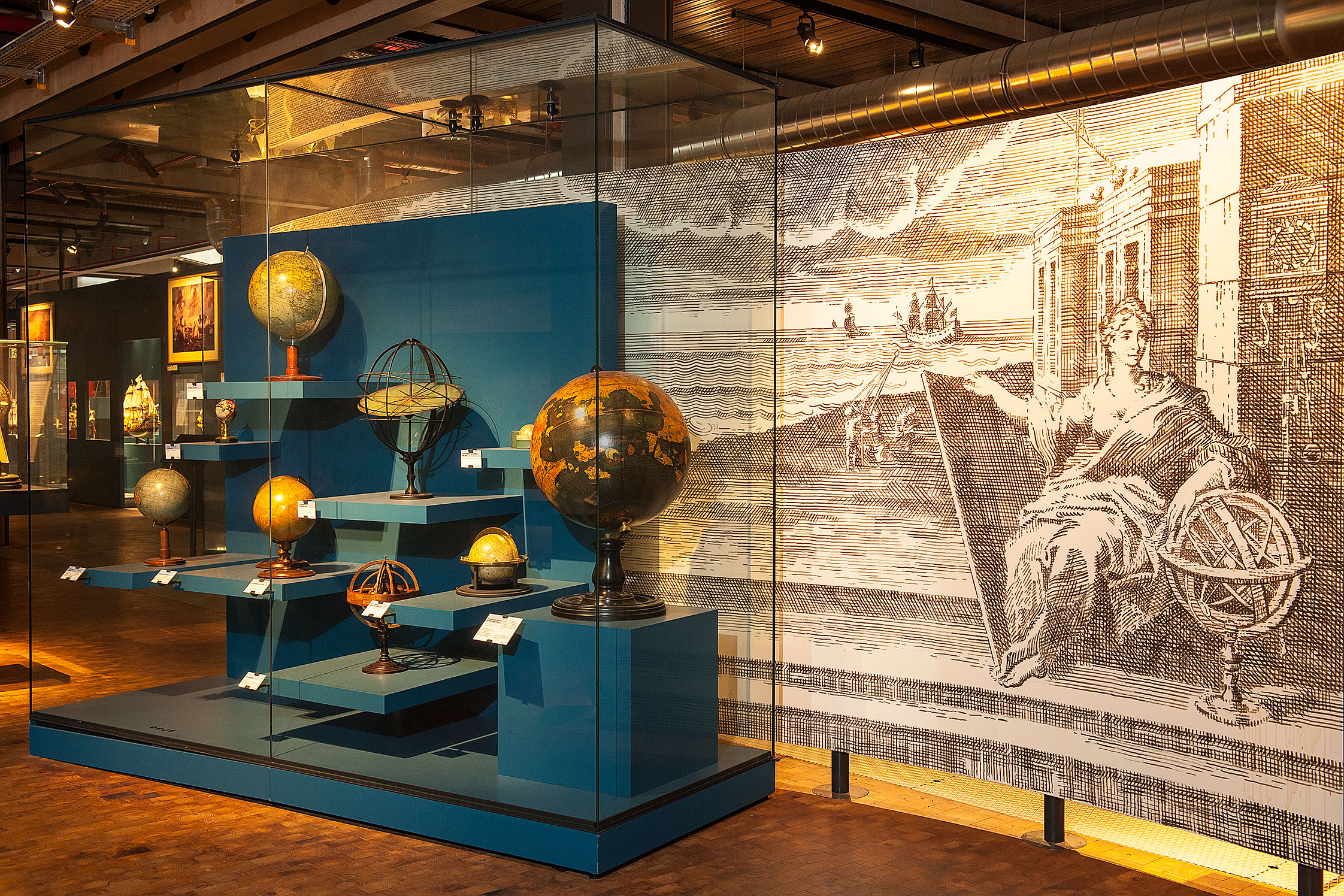 Vor einem großen, schwarz-weißen Wandbild stehen in einer Vitrine mit blauen Sockeln verschiedene historische Globen.